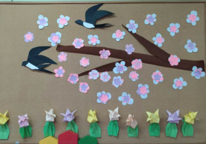 Tablica korkowa. Na tablicy przyczepione są kolorowe tulipany origami . Z prawej stronie wykonana z bibuły i papieru gałązka z półprzestrzennymi kwiatami jabłoni. Pomiędzy gałązkami latają trójwymiarowe jaskółki z papieru.