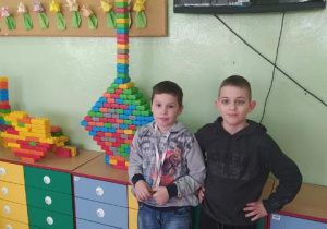Dwóch chłopców stoi przy kolorowych szafkach na których ustawili swoją wieżę z kolorowych klocków. W tle na ścianie wisi telewizor i korkowa tablica.