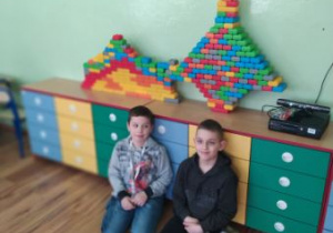 Dwóch chłopców siedzi na podłodze przed kolorowymi szafkami. Na szafce stoi wieża z kolorowych klocków.