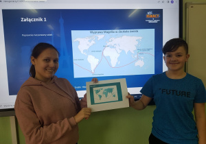 Chłopiec i dziewczyna stoją na tle tablicy interaktywnej na której wyświetlona jest mapa kontynentów. Uczniowie trzymają razem kartkę papieru .