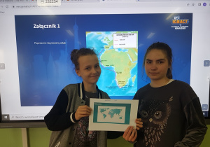 Dwie uczennice stoją na tle tablicy interaktywnej na której wyświetlona jest mapa kontynentów. Uczniowie trzymają razem kartkę papieru .