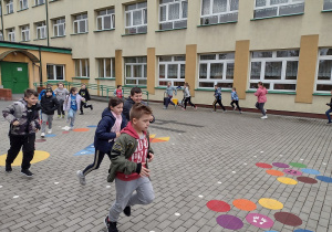 Uczniowie ćwiczą swobodny bieg w kółku na placu szkolnym. W tle budynek szkoły