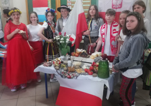Na pierwszym planie stoi stolik przykryty biało-czerwonym materiałem. Na stoliku stoją potrawy związane z Polską. Za stolikiem stoją uczniowie. Para w strojach w strojach ludowych , harcerka z flagą Polski i kibic. W tle biało czerwona flaga.