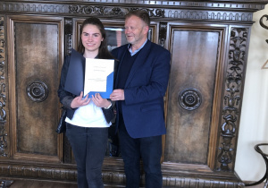 Laureatka konkursu polonistycznego trzyma dyplom który wręcza jej Dyrektor Delegatury Kuratorium Łódzkiego.W tle zdobiona szafa.