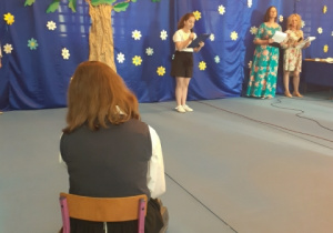 Uczennica siedzi na krześle odwrócona plecami. W tle scena na której stoi dziewczynka iz prawej strony stoja obok siebie dwie kobiety.