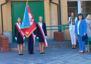 Poczet sztandarowy SP14 . Chłopiec trzyma sztandar szkoły. Po jego bokach stoja uczennice ubrane w strój galowy. Z prawej strony stoi Pani Dyrektor. W tle budynek szkoły