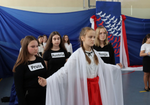 Sala gimnastyczna. Na pierwszym planie stoi dziewczyna ubrana na biało - czerwono. Za nią stoją uczennice ubrane na czarno z napisami Rosja, Prusy i Austria.Tło niebieskie.
