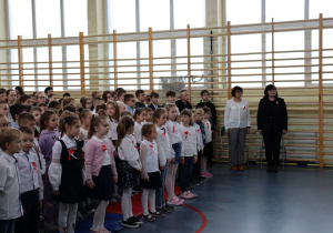 Sala gimnastyczna. Duża grupa dzieci stoi na baczność. Wszyscy są w strojach galowych. W tle drabinki gimnastyczne.