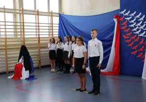 Sala gimnastyczna. Uczniowie w strojach galowych śpiewają.Niebieskie tło.