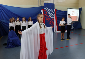 Sala gimnastyczna. Na pierwszym planie stoi dziewczyna ubtana na biało- czerwono. Za nią stoja uczniowie ubrani na galowo. Tło niebieskie.