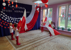 Wnętrze sali przedszkolnej. Czterech chłopców macha flagami. W tle dekoracja z flagą i okna.