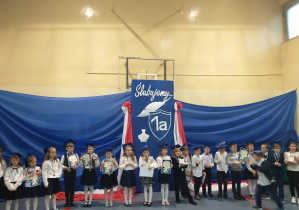 Sala gimnastyczna.Uczniowie ubrani w stoje galowe i biirety stoja na niebieskim tle.
