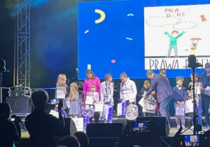 Grupa dzieci z torbami prezentowymi stoi na scenie . W tle wyświetlona praca plastyczna dziecka. Z lewej strony stoi prezenterka ubrana na biało.