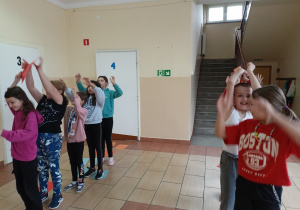 Korytarz szkolny. Dzieci ubrane na sportowo stoją w dwóch rzędach. Uczniowie podają sobie nad głowami kartkę papieru. W tle klatka schodowa.