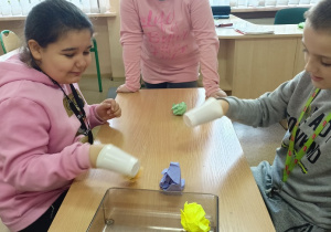 Wnętrze klasy.Trzech uczniów siedzi przy ławce. Na ławce stoi pojemnik ze zgniecionymi kulami papieru. Uczniowie mają założone na dłonie plastikowe kubki.