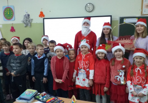 Wnętrze klasy. Na tle tablicy interaktywnej stoi Mikołaj i grupa uczniów młodszych w czerwonych czapkach.