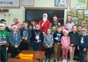 Wnętrze klasy. Na tle tablicy interaktywnej stoi Mikołaj i grupa uczniów młodszych w czerwonych czapkach.
