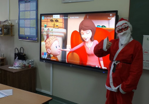 Osoba przebrana za Świętego Mikołaja stoi na tle tablicy interaktywnej..