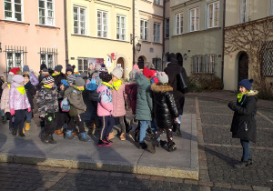 Du za grupa dzieci ubranych w kurtki stoi na wybrukowanym placu . W tle kamienice