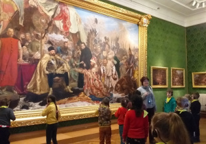 Wnętrze zamku. Grupa dzieci stoi przed wielkim obrazem. W tle zielona ściana.