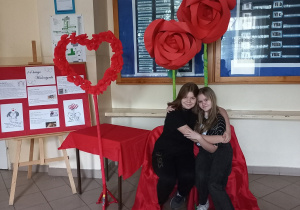 Na pierwszym planie na krzesłach przykrytych czerwonym materiałem siedzą dwie dziewczyny ubrane na czarno. Obok stoi stolik przykryty czerwonym materiałem. Za stolikiem na stojaku umieszczona jest obręcz w kształcie serca. W tle gablota szkolna i olbrzymie czerwone róże z papieru