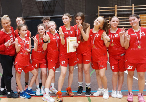 Hala sportowa. Na boisku w szeregu stoi grupa dziewcząt w czerwonych strojach sportowych. Po lewej stronie stoi trenerka. Wtle drabinki gimnastyczne.