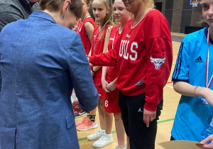 Hala sportowa. Hala sportowa. Na boisku w szeregu stoi grupa dziewcząt w czerwonych strojach sportowych. Przed nimi tyłem stoi kobieta. Kobieta odznacza jedną z dziewcząt medalem. Obok kobiety stoi mężczyzna.