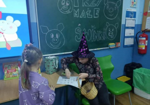 Na tle zwykłej tablicy i tablicy interaktywnej siedzi przy stoliku dziewczynka przebrana za czarownicę. Obok niej tylem stoi druga dziewczynka.