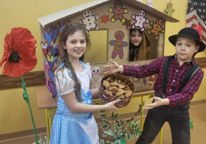 Na tle piernikowej chatki stoi dziewczynka i chłopiec grający rolę Jasia i Małgosi . Dziewczynka trzyma w ręku koszyk z piernikami. Z tyłu za chatką na ścianie wisza tablice edukacyjne.
