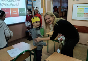 Mały chłopiec w koronie stoi przed pochyloną nad nim uśmiechniętą kobietą. W tle tablica interaktywna i otwarte drzwi. Z tylu za chłopcem stoją trzy dziewczynki.