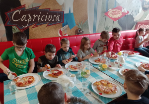 W kolorowym wnętrzu stoi stół. Po obydwu stronach stołu siedzą chłopcy. Na stole stoją szklanki i dzbanek z wodą. Przed chłopcami stoją talerze z pizzą. Chłopcy jedzą pizzę.