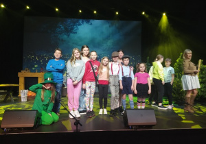 Ciemne tło sceny. U góry rampa ze światłami. Na środku stoi grupka uczniów. Dziecko z lewej strony siedzi na podłodze.Po prawej stronie, bokiem stoi kobieta.