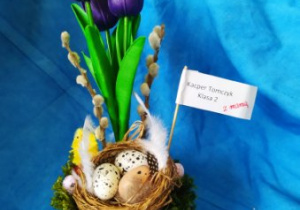 Niebieskie tło. Stroik wielkanocny kwiatowy umieszczony w ceramicznym naczyniu w kształcie skorupki od jajka.
