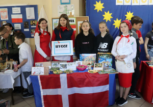 Na pierwszym planie stoi stolik z flagą Danii. Na stoliku stoją potrawy. Za stolikiem stoją uczennice ubrane w biało-czerwone stroje.