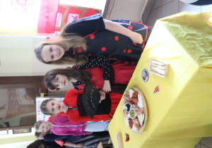 Na pierwszym planie stoi żółty stolik z potrawami. Za stolikiem stoją trzy dziewczyny ubrane w czerwono-czarne stroje. W tle flaga Hiszpanii.