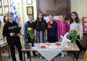 Na stoliku stoją potrawy i wazony z tulipanami . Za stolikiem stoją uczniowie. W tle flaga Hiszpanii.