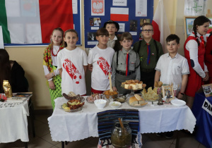 Na stoliku stoją potrawy. Przed stolikiem stoi słoik z ogórkami. Za stolikiem stoją uczniowie ubrani w koszulki z symbolami Polski. W tle flaga Polski.