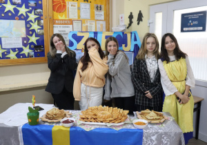 Na stoliku stoją potrawy. Z przodu stołu zamocowana jest flaga Szwecji. Za stołem stoją uczennice. W tle napis Szwecja i gazetki szkolne.