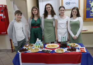 Na stoliku przykrytym czerwonym płótnem stoją potrawy. Do stolika przymocowano flagę Węgier. Za stołem stoją dziewczęta. W tle gazetka szkolna.