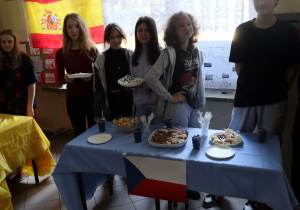 Na stoliku stoją potrawy. Do stolika przymocowana jest flaga Czech. Za stolikiem stoi grupa uczniów.