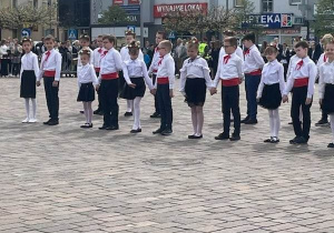 Plac. Na placu duża grupa uczniów ubrana w biało -czarne stroje. W tle kamienice.