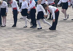 Plac. Na placu duża grupa uczniów ubrana w biało -czarne stroje. W tle kamienice.