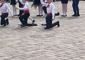 Plac. Na placu duża grupa uczniów ubrana w biało -czarne stroje. Uczniowie tańczą .W tle kamienice.