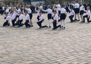 Plac. Na placu duża grupa uczniów ubrana w biało -czarne stroje. Uczniowie tańczą.W tle kamienice.