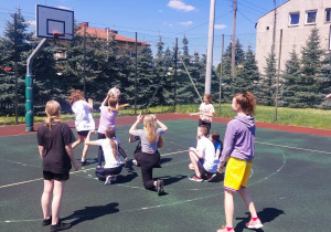 Boisko szkolne ogrodzone siatką. Na boisku grupa dziewcząt i chłopców gra w piłkę koszykową. W tle wysoka kamienica i drzewa iglaste.