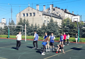 Boisko szkolne ogrodzone siatką. Na boisku grupa dziewcząt i chłopców gra w piłkę nożną. W tle wysoka kamienica.