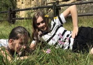 Dwie dziewczyny lezą na trawie. Za nimi ogrodzenie z belek i wapienna ściana grot.