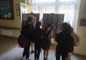 Korytarz szkolny. Grupa uczennic stoi przed sztalugami. Na sztalugach umieszczono tablice korkowe ze zdjęciami oczu. W tle okno.