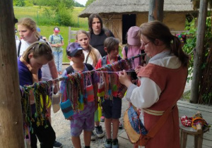 Przed chatą w średniowiecznym stroju stoi kobieta. Kobieta pokazuje dzieciom różne tkackie węzły. W tle drewniana chata.