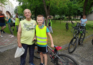 Mężczyzna stoi z chłopcem. Chłopiec w kamizelce odblaskowej opiera się o rower. Po prawej stronie stoją rowery.Po lewej stronie stoi grupa osób. W tle drzewa.